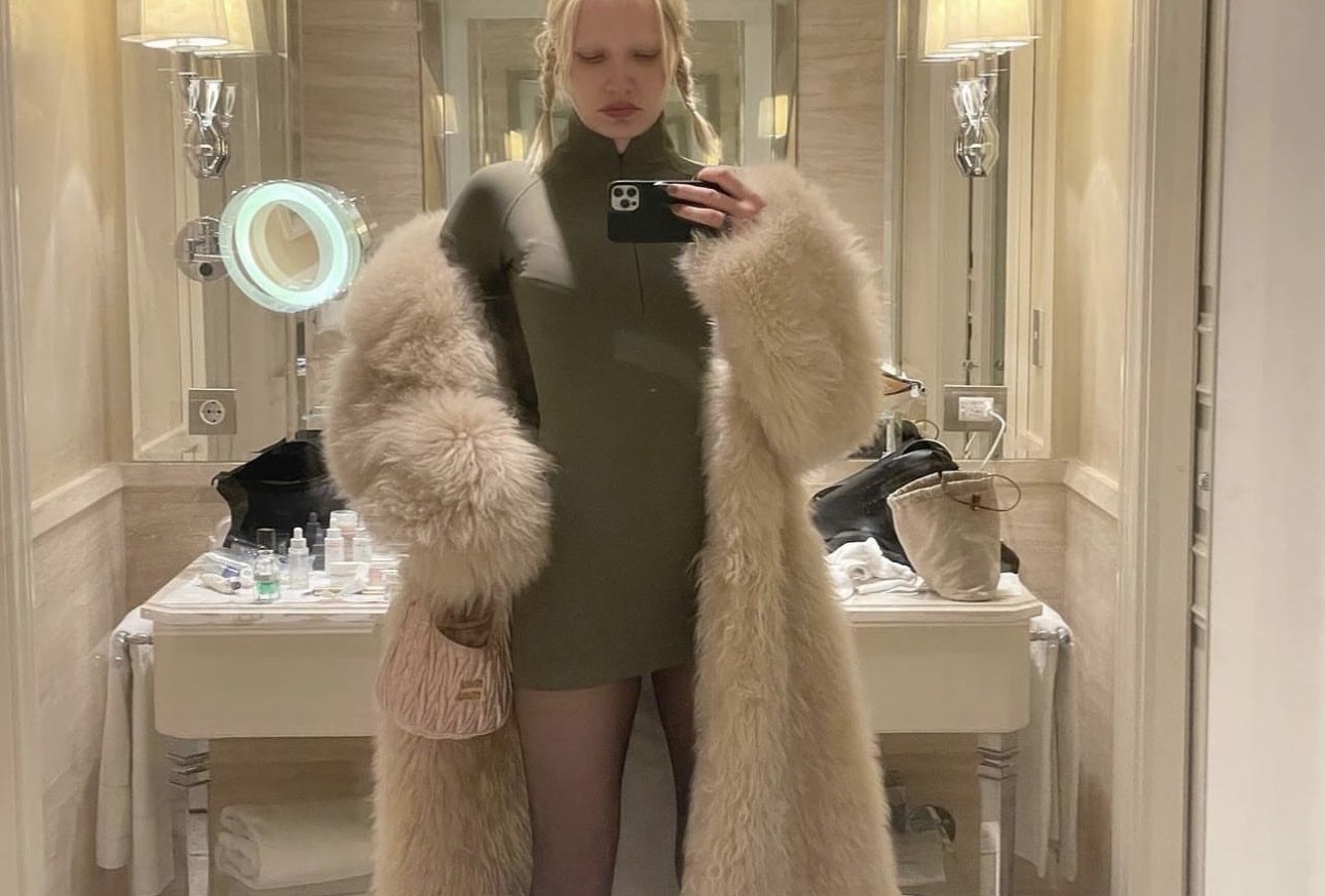 Mirror photograph of a person styling a textured coat over a fitted dress